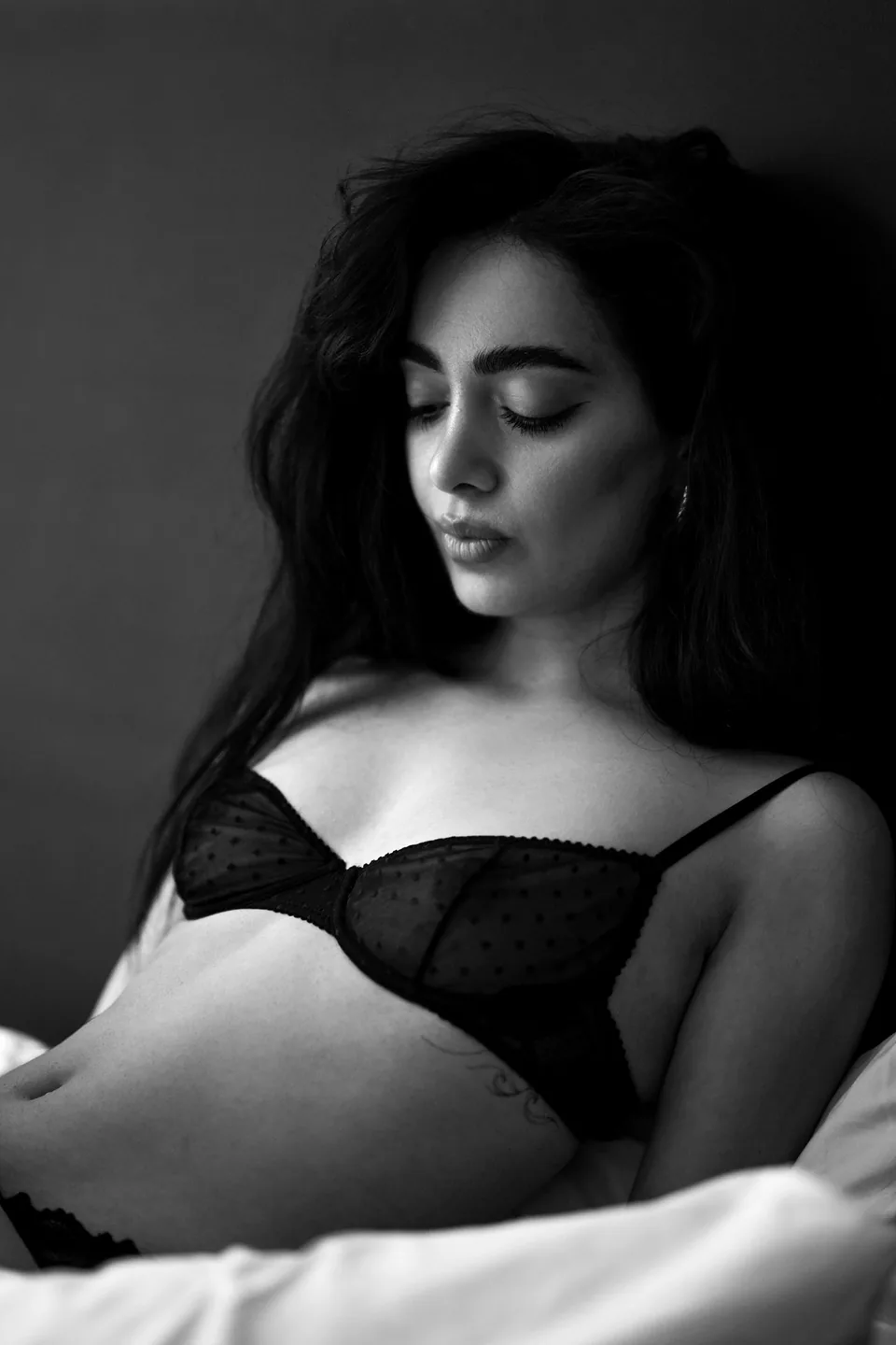 Woman with long dark hair in a sheer black bra, eyes lowered, in soft window light